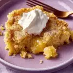 Plated serving of lemon dump cake with poppy seeds topped with whipped cream on a pastel purple plate