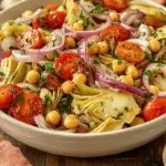 Close-up of Tuscan artichoke tomato salad with cherry tomatoes, chickpeas, red onions, herbs, and vinaigrette in a white bowl on a rustic wood surface